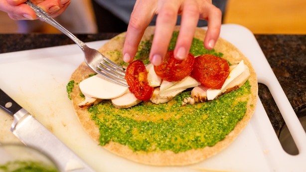 Close-up of hands assembling flatbread with pesto, fresh mozzarella slices, roasted tomato, and grilled chicken on a cutting board.