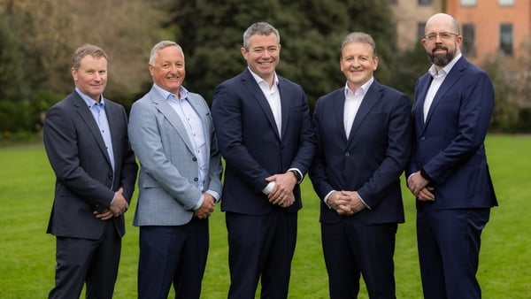 Five businessmen in suits pose for a photo in a garden