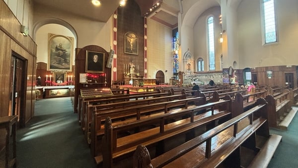 Several brown benches can be seen inside a church. The windows are colourful stained glass.