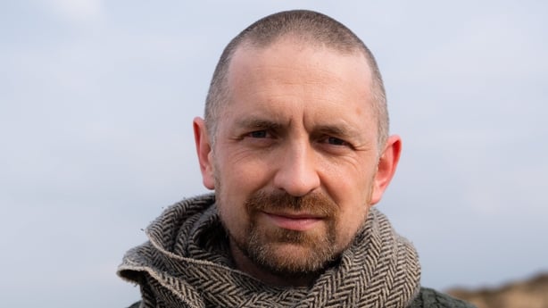 A close up headshot of Billy Mag Fhloinn looking straight into the camera. He is outside with the sky behind him