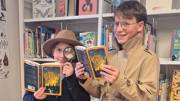 Two children, one wearing a hat and holding a magnifying glass, hold copies of a book with bookshelves in behind them