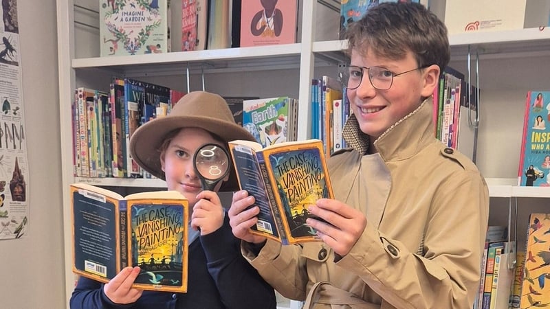 Two children, one wearing a hat and holding a magnifying glass, hold copies of a book with bookshelves in behind them