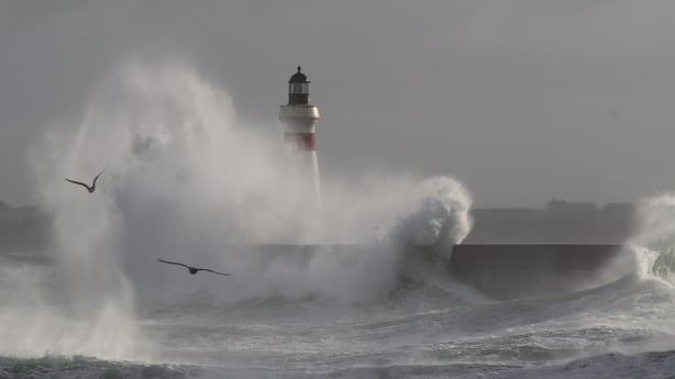 Big waves break against a pier with a lighthouse breakwater with beacon and seagulls