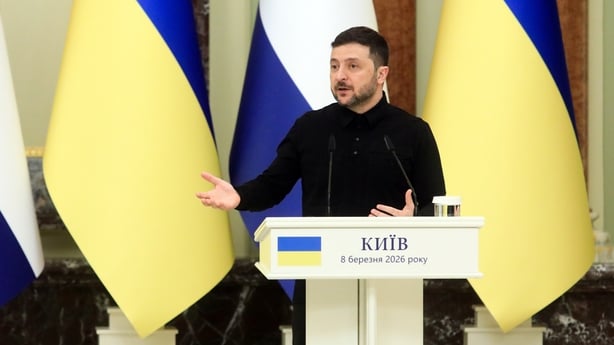 Volodymyr Zelensky gestures as he speaks to the media with two ukranian flags behind him