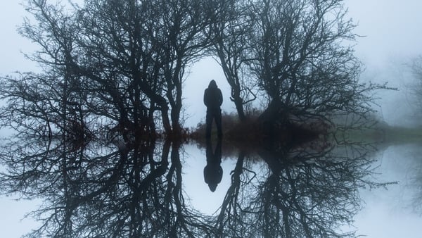 A creepy reflection upside down edit, of a hooded figure, standing in an archway of trees on a dark, eerie winters day in the countryside