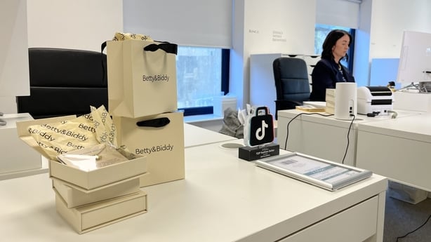 Woman sitting at a desk with Betty and Biddy bags on another desk in the foreground