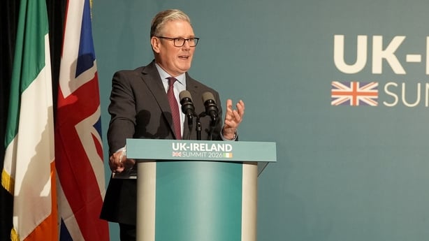 A photo of british prime minister keir starmer speaking at a lectern with irish and uk flags hanging behind him