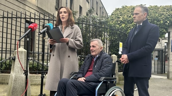 a woman stands before microphones reading from a statement with a man in a wheelchair and another man standing beside him