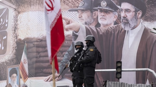 Two armed police officers stand guard in front of a giant banner depicting a portrait of Iran's new Supreme Leader, Ayatollah Mojtaba Khamenei, and military commanders