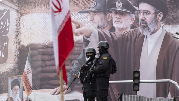 Two armed police officers stand guard in front of a giant banner depicting a portrait of Iran's new Supreme Leader, Ayatollah Mojtaba Khamenei, and military commanders