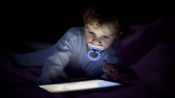 A small child lies on a bed with a soother, intently focused on a glowing tablet screen in the darkness 
