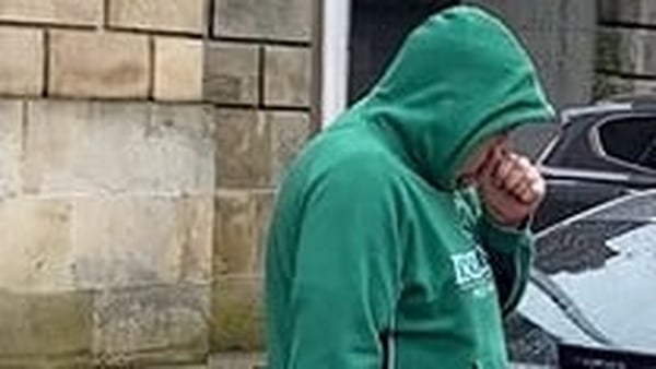 a man wearing a green hoodie with the hood up puts his hand to his face