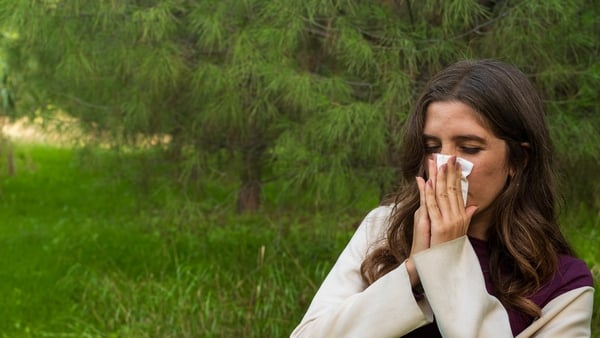 A caucasian woman suffering from a cold uses a tissue to blow her nose while standing in a green natural park environment — captured in soft daylight, conveying stage 1 runny nose symptoms.