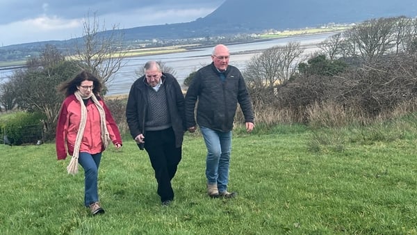 Two men and a woman walk through a field with bushes and the sea behind them
