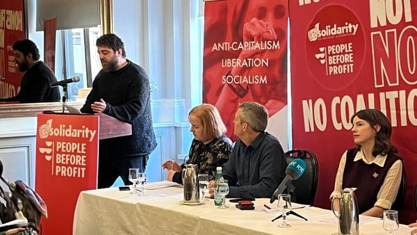three people sit at a table listening as another person speaks at a lectern with signs behind them