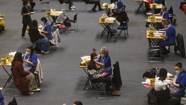 A large area has multiple tables and chairs as people wearing facemasks receive a vaccination