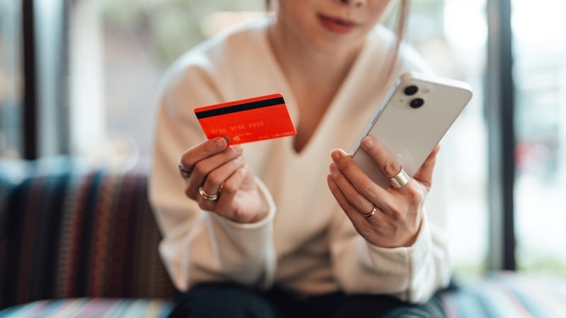 Midsection of young Asian woman holding a credit card while using smartphone, sitting on sofa. Online shopping and electronic payment.