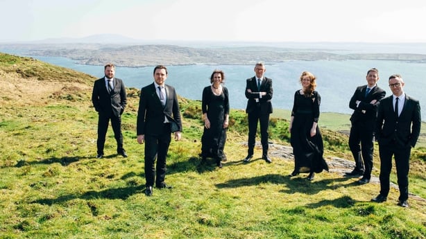 The vocal group Resurgam stand on a grassy cliff with the sea behind them on a sunny day. There are 5 men and 2 women wearing black and standing in a staggered line.