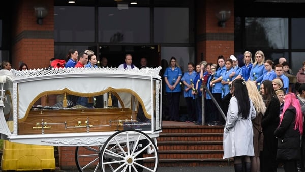 People, including healthcare workers in uniform, watch on as a horse drawn hearse, carrying a coffin, arrives at a church