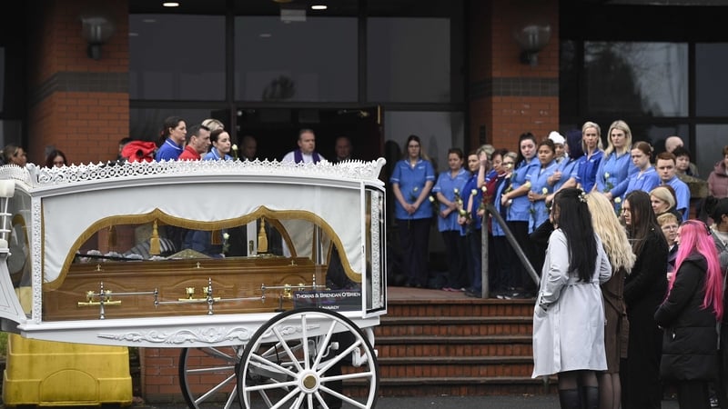 People, including healthcare workers in uniform, watch on as a horse drawn hearse, carrying a coffin, arrives at a church