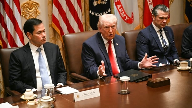 This photographs shows from left Marco Rubio, Donald Trump and Pete Hegseth sitting at a table with name plaques in front of them and flags hanging behind them