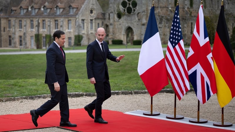 Two men wearing suits - Marco Rubio and Jean-Noel Barrot - walk on a red carpet with flags hanging on flagpoles