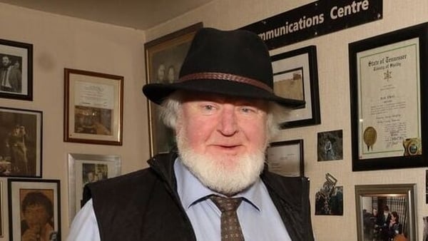 Proinsias O Rathaille, in hat, with walls hung with many pictures behind him