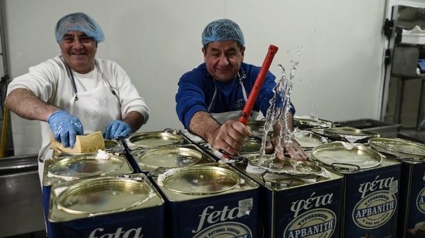 Two people wearing blue net head coverings and aprons smile as the work putting feta into containers