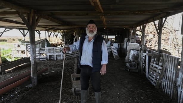 A man holding a long stick poses for a photograph on a farm in front of empty animal pens