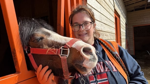 A woman smiles as she poses beside a horse who's head sticks out of a stable door