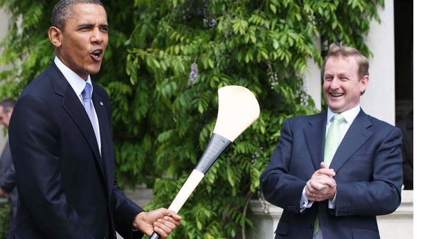 Barack Obama and Enda Kenny stand outdoors with a tree in the background. Obama is holding a hurley.