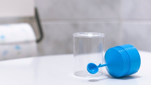 A stool sample container for fecal tests in the bathroom with a roll of toilet paper in the background.