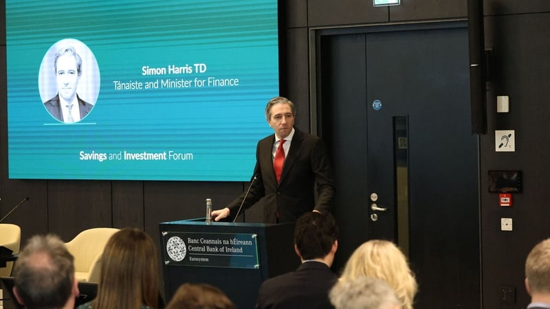 Simon Harris stands at a lectern with a large screen showing his photo and name behind him