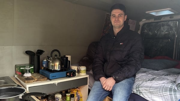Conor Davidson sitting on a bed in his van with a kettle and other kitchen items on a tabletop beside him