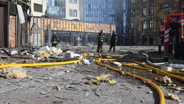 Fire hoses and firefighters stand in an area of damaged buildings