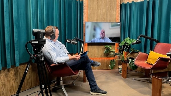 A man sits in a studio talking to a person remotely, who's image is on a big screen