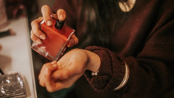 The image shows a young woman applying perfume to her wrist, capturing a delicate and intimate moment associated with personal care, beauty routines, and self-expression. The gesture highlights elegance, femininity, and sensory experience, commonly used t