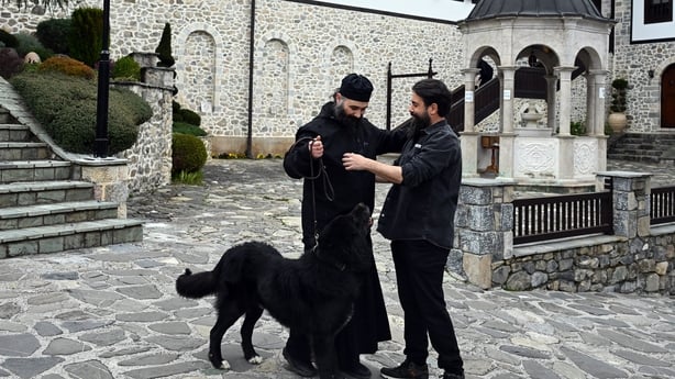 Two men chat as a large black dog stands beside them 