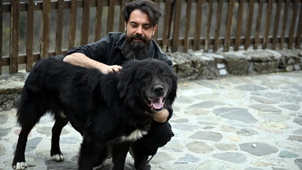 A man squats on the ground beside a large black dog
