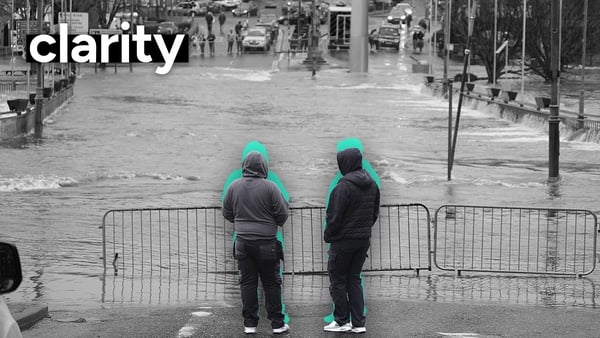 Monochrome image ot two people wearing jackets with hoods up standing at a metal barrier beyond which lies a flooded street