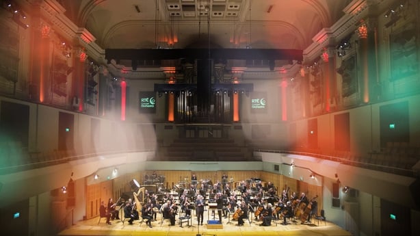 The full RTÉ Concert Orchestra are on the stage of the National Concert Hall seated with their instruments. The photo is taken from the back of the hall, showing a wide shot of the stage, balcony and ceiling 