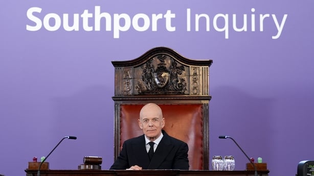 A photograph of adrian fulford sitting at a desk with a sign reading 'southport inquiry' behind him