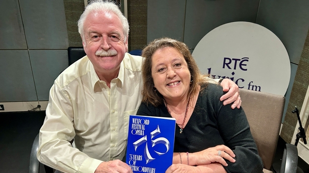 Marty Whelan sits beside Rosetta Cucchi with one arm around her. They are smiling at the camera and holding up a blue programme for Wexford Festival Opera.