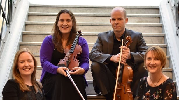 The four members of the Ficino Quartet sit on a set of stairs - two on the top and two below them at either side. They are holding their string instruments and smiling at the camera