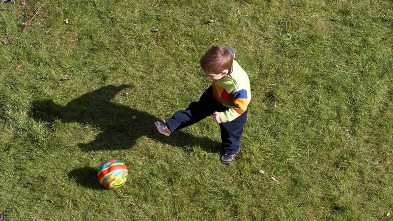 Small young boy outside on a sunny garden lawn playing with and kicking a football