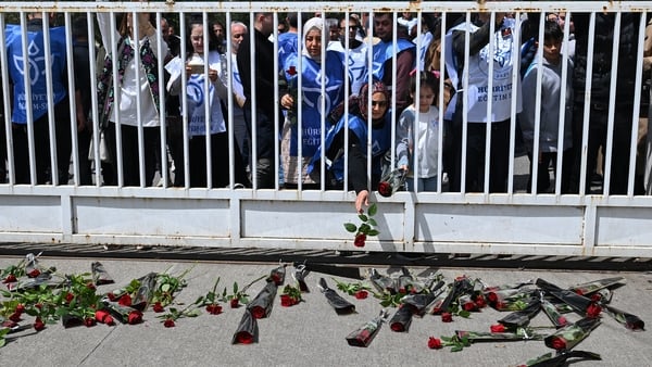 Image shows dozens of teenagers standing behind a white fence with flowers laid on the ground.