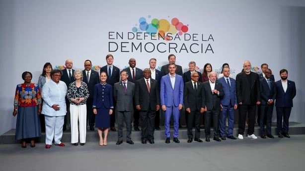 A group of people pose for a photograph with a sign reading 'en defnsa de la democracia' in the background