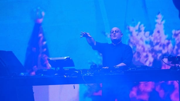 a man in clerical garb gestures as he stands behind sound desks