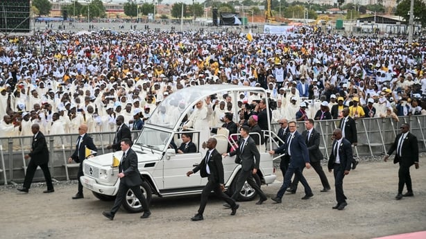 Pope Leo waves as security guards walk beside the popemobile as thousands watch on in Kalimba, Angola