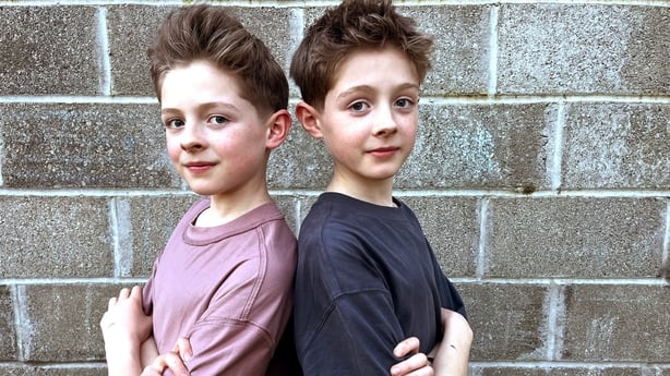 Twin brothers Max and Sonny pose back to back in front of a grey wall.